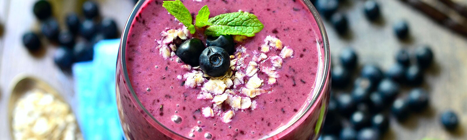 Center frame is a smoothie in a glass topped with blueberries and a mint leaf. Out of focus in the background blueberries and mint sit on a table. Center frame is a smoothie in a glass topped with blueberries and a mint leaf. Out of focus in the background blueberries and mint sit on a table.
