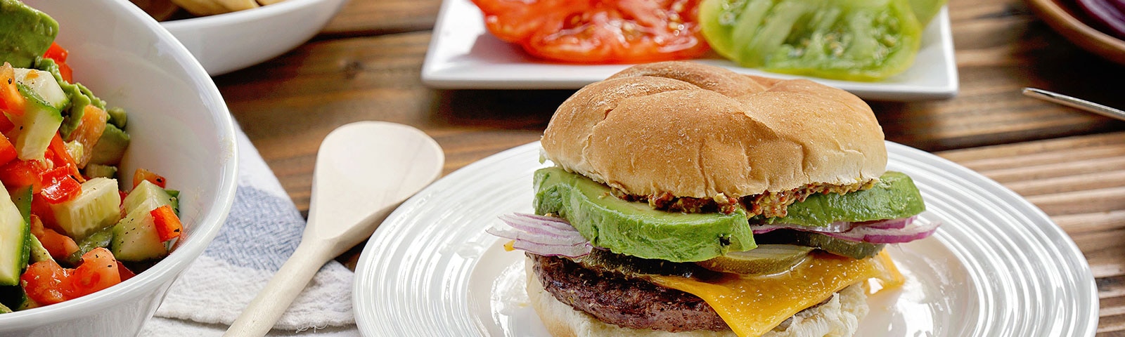 A burger loaded with toppings sits on a white plate surrounded by various burger topping ingredients