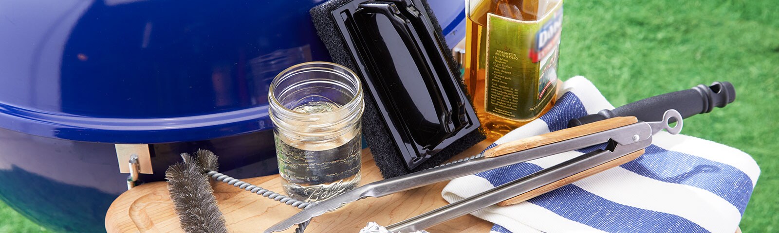 Blue outdoor drill with an attached shelf, on which sits a cloth, cooking oil, tongs, a wire brush, tin foil, scrub brush and mason jar.