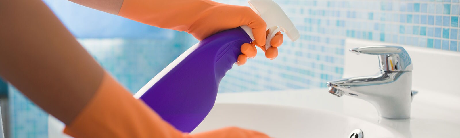 Two arms wearing rubber gloves cleaning a bathroom sink, spray bottle in the left hand with a cloth to wipe in the right