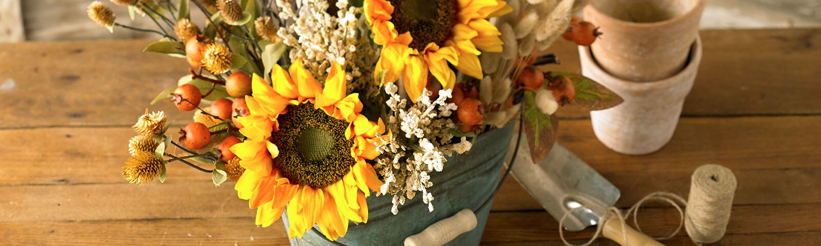 Sitting on a rustic wooden table a metal bucket is filled with sunflowers and other fall blooms.