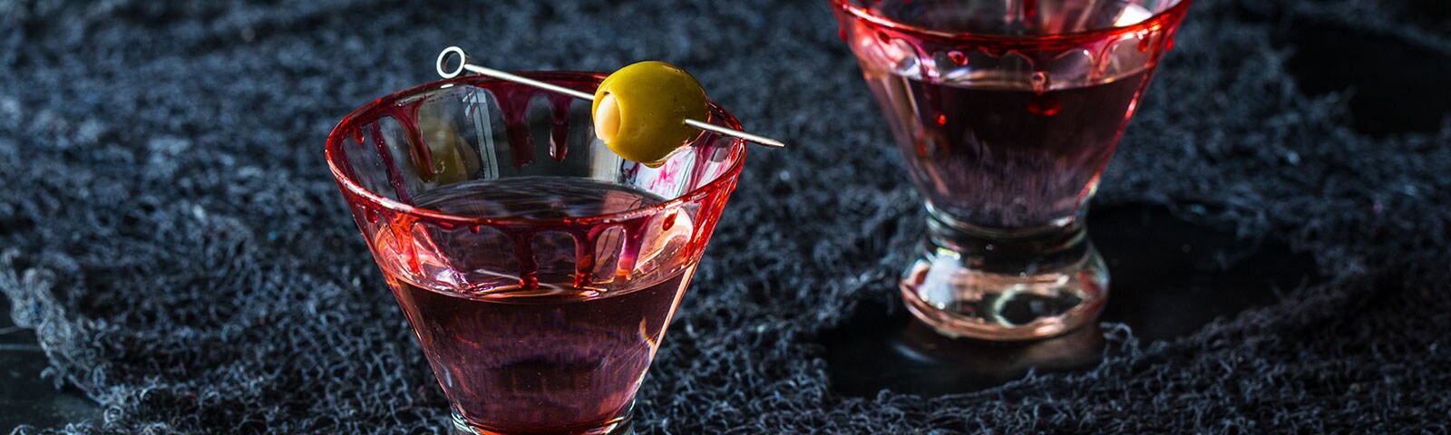 Two cocktail glasses with red liquid dripping down their sides sit on a dark blue cloth