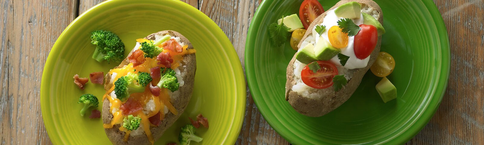 Two baked potatoes sitting on separate plates on a wooden table. Each potato is loaded with various toppings from bacon, to broccoli, to avocado. Two baked potatoes sitting on separate plates on a wooden table. Each potato is loaded with various toppings from bacon, to broccoli, to avocado.