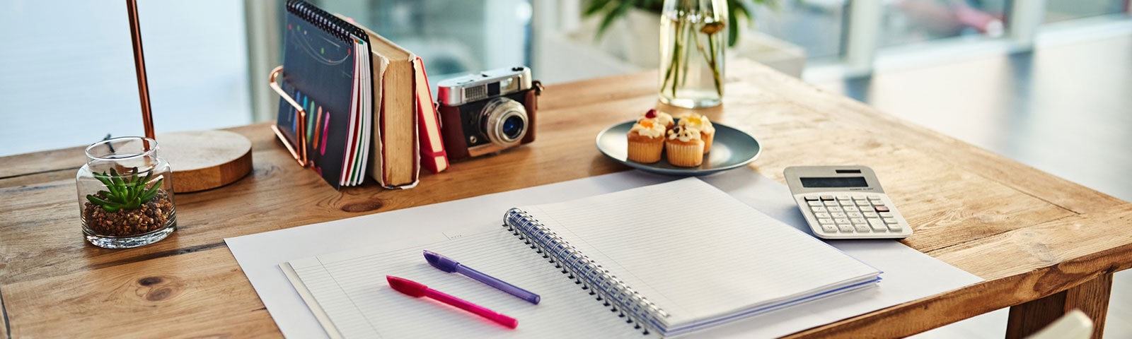 Pen and paper on wooden desk with cupcakes and plant Pen and paper on wooden desk with cupcakes and plant